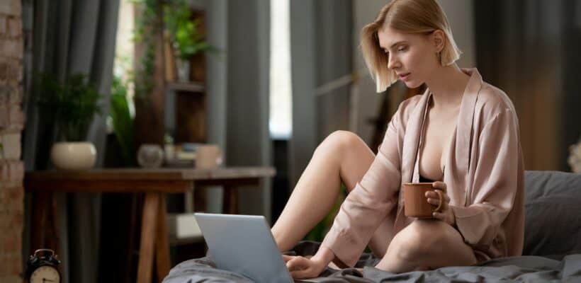 Blond pretty woman in black lingerie and beige silk robe having coffee on bed